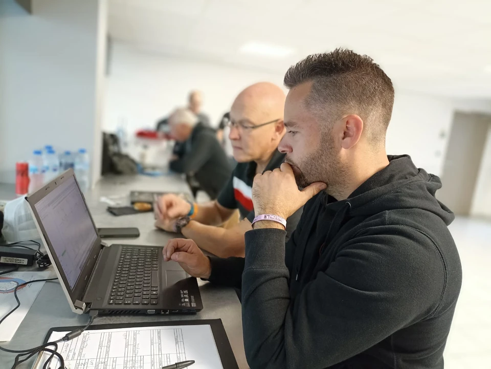 Pilote en tenue de compétition (combinaison, casque intégral) au volant d'une Porsche de sport ou de compétition (rouge, noir ou couleur d'équipe), captured en action sur piste de circuit avec virages et marquages visibles, montrant la concentration et l'expertise du conducteur. Image de qualité professionnelle, prise de côté ou de trois-quarts pour bien voir le conducteur et la voiture en dynamique.